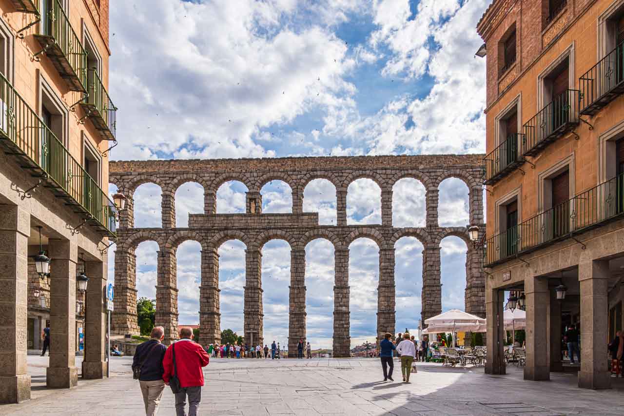 People are in a square that is dominated by a multi-arched, two-tiered stone aqueduct built by the ancient Romans.