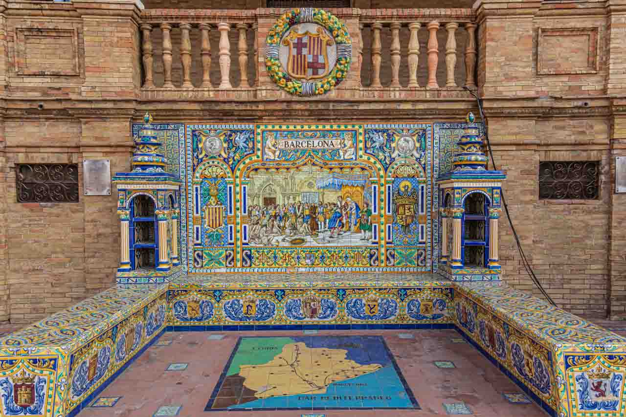A multi-coloured three-sided tiled bench with a tiles mural against a brick wall.