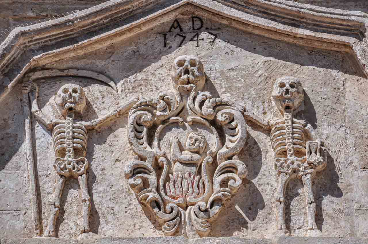 A skull and two skeletons carved into stone. One skeleton is holding an hourglass, and the other skeleton is holding a scythe.