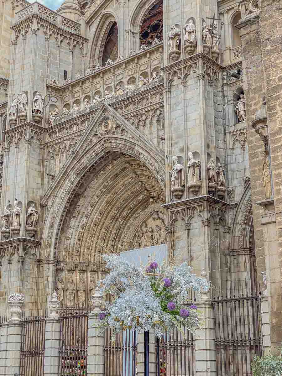 The facade of a gothic cathedral with white and purple flowers on a pole in front of the cathedral.