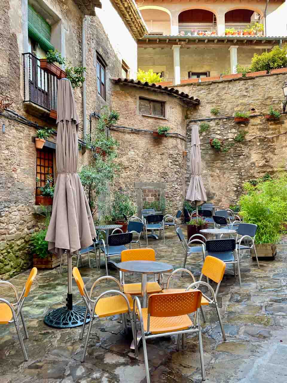 Cafe tables and chairs and umbrellas are set up in a small square in fromt of stone buildings.