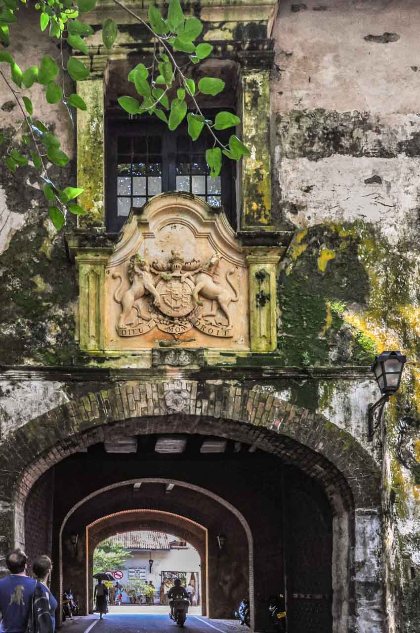 A short stone tunnel with with a motor bike rider and lady inside it ands two men viewing the British Coat of Arms on the facade of the tunnel.