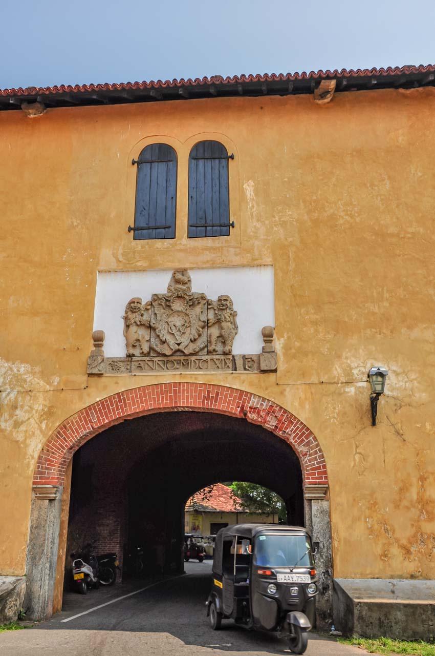 A yellow wall with a tunnel and a tuk tuk coming through it. The Dutch East Indian Company crest is on the facade above the tunnel.