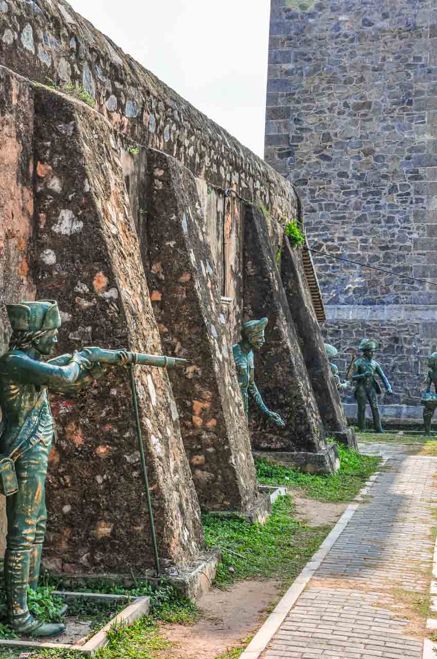 Statues of men with firearms are placed between the bastions on a stone wall.